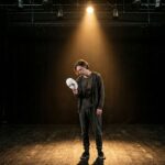 Ator em um palco escuro sob um único holofote, segurando uma máscara branca afastada do rosto, com expressão de cansaço e alívio.