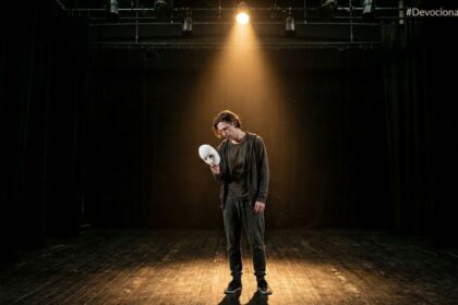 Ator em um palco escuro sob um único holofote, segurando uma máscara branca afastada do rosto, com expressão de cansaço e alívio.