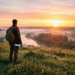 Homem em pé numa colina verde segurando um projeto enrolado, olhando para o horizonte com um nascer do sol sobre um vale e um rio.