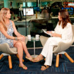 Fotografia em plano médio da atriz Luana Piovani. Ela é uma mulher branca, com cabelos loiros presos de forma despojada e veste uma camiseta de malha na cor cinza. Luana tem uma expressão facial séria e reflexiva, com o olhar direcionado ligeiramente para o lado. O fundo da imagem é um ambiente interno desfocado, em tons escuros e amarelados, que destaca o rosto da atriz em primeiro plano.