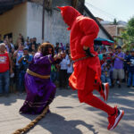 Ator fantaseado de Jesus segurando um crucifixo e apontando para o outro ator, fantasiado de Diabo.