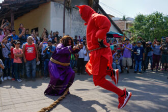 Ator fantaseado de Jesus segurando um crucifixo e apontando para o outro ator, fantasiado de Diabo.